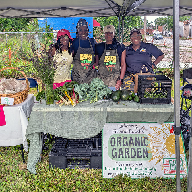 ETC team members smiling behind a produce table with kale, Swiss chard, and zucchini at the Kingsway East Farmers Market